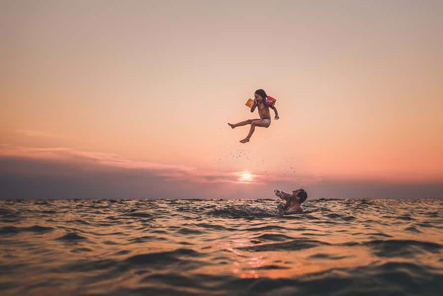 Criança saltando das ondas do oceano ao pôr do sol com outra criança nadando abaixo. — Filmes Papéis de Parede