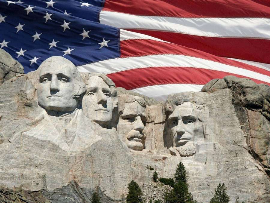 Mount Rushmore mit gemeißelten Präsidentengesichtern und einer großen amerikanischen Flagge, die im Hintergrund weht. — Flagge Der USA Wallpaper