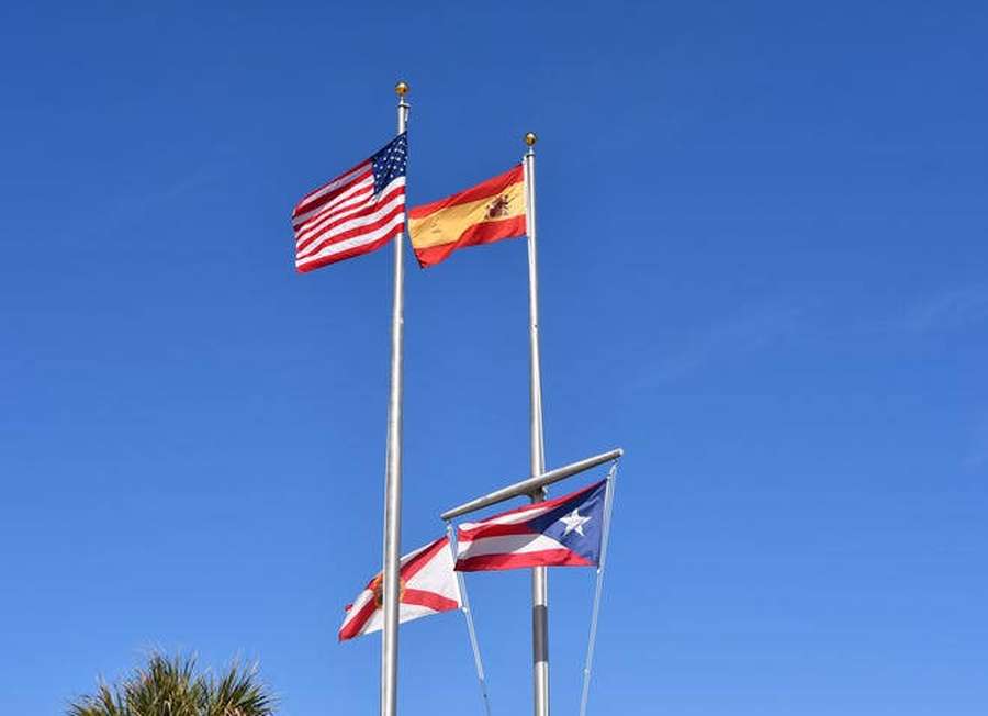Drei Nationalflaggen wehen an Stangen vor einem klaren blauen Himmel mit einer sichtbaren Palme. — Flagge Spaniens Wallpaper