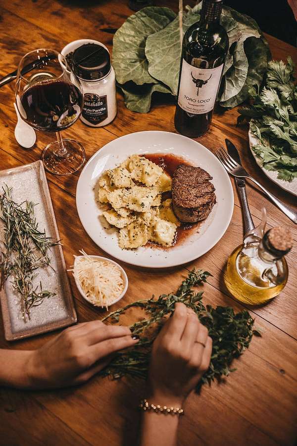 Hände arrangieren frische Kräuter neben einem angerichteten Steak und Gnocchi mit Wein und Olivenöl auf einem Holztisch. — Fleisch Wallpaper