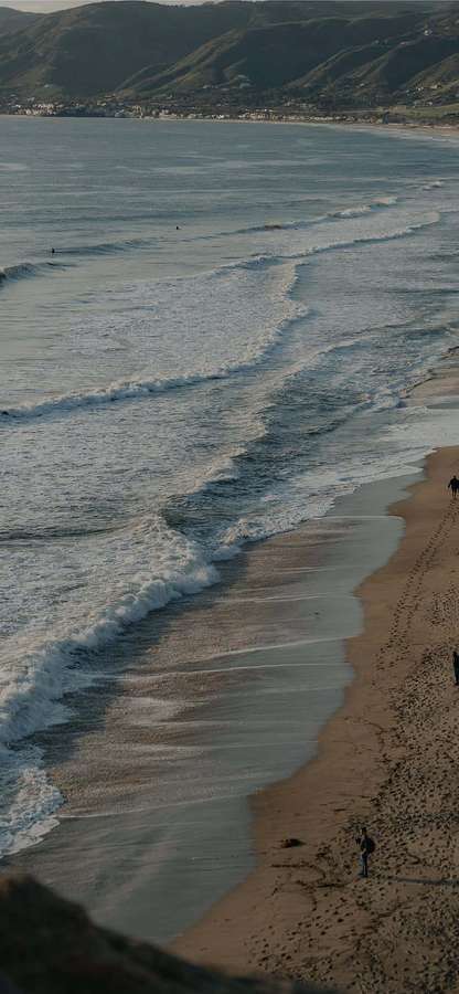 Las olas ruedan en una playa de arena con huellas que conducen a colinas lejanas. — Fondods de Android Malibu Fondos