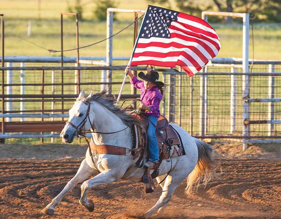 Joven montando caballo blanco ondeando bandera estadounidense en arena de rodeo. — Fondos de Rodeo Fondos