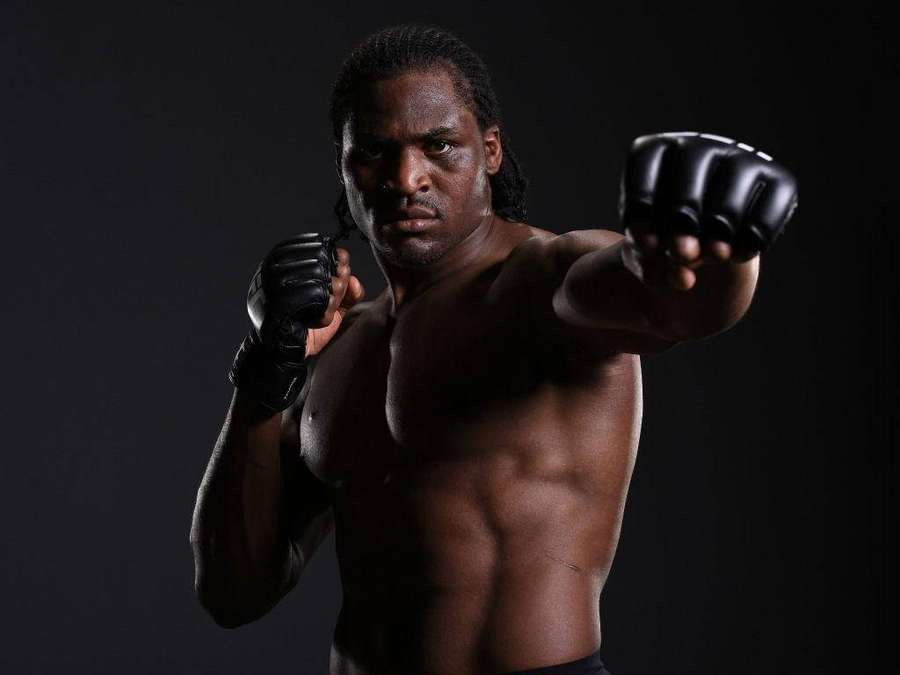 A shirtless male fighter in black gloves poses intensely in a dark studio. — Francis Ngannou Background