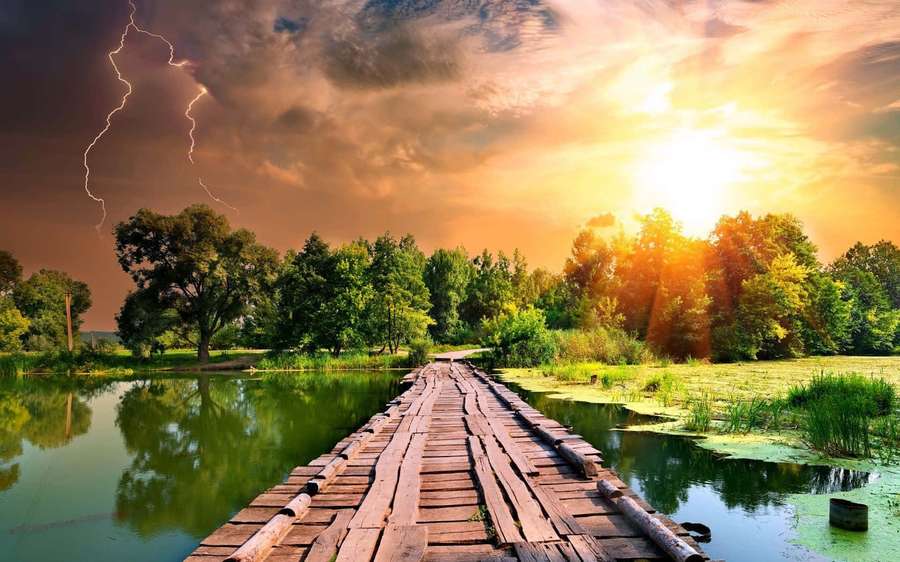A rustic wooden bridge crosses a pond surrounded by trees under a stormy sunset with lightning. — Free Background