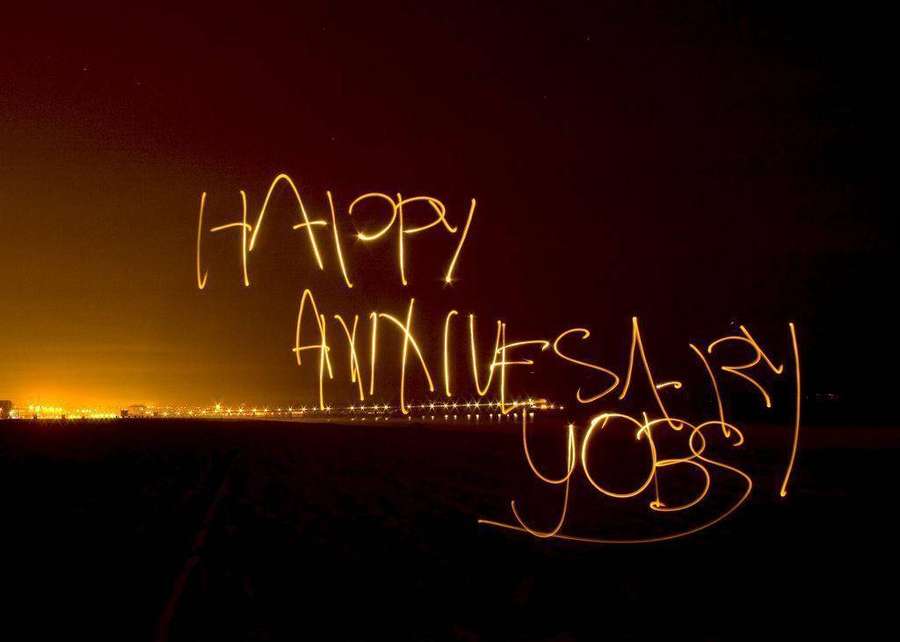 Happy anniversary geschrieben in leuchtenden Lichtspuren über einer nächtlichen Stadtlandschaft. — Frohes Jubiläum Wallpaper