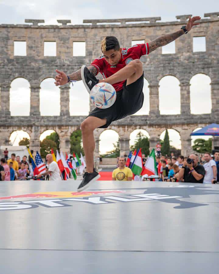 Futbolista ejecutando un truco en el aire en un evento de Red Bull frente a un anfiteatro antiguo. — Fútbol Freestyle Fondos de pantalla