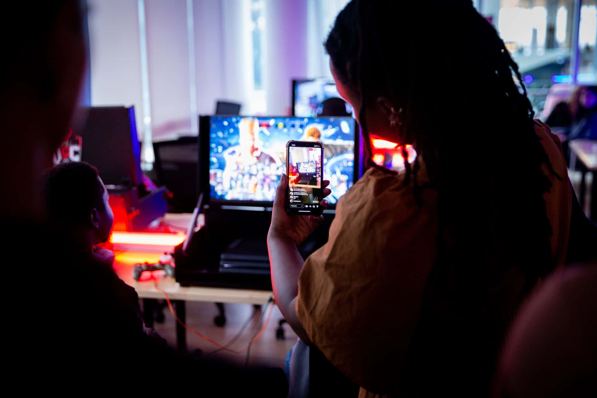 Person tar opp en videospilløkt på telefon i mørkt rom med lysende skjermer. — Game Development Bakgrunner