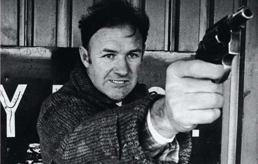 Man in textured coat aiming revolver with tense grin in gritty urban setting — Gene Hackman Wallpaper