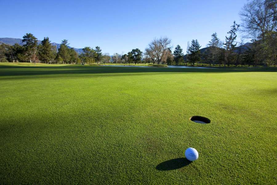 Golf ball near hole on vibrant green fairway with trees and mountains in background. — Golf Course Desktop Wallpaper