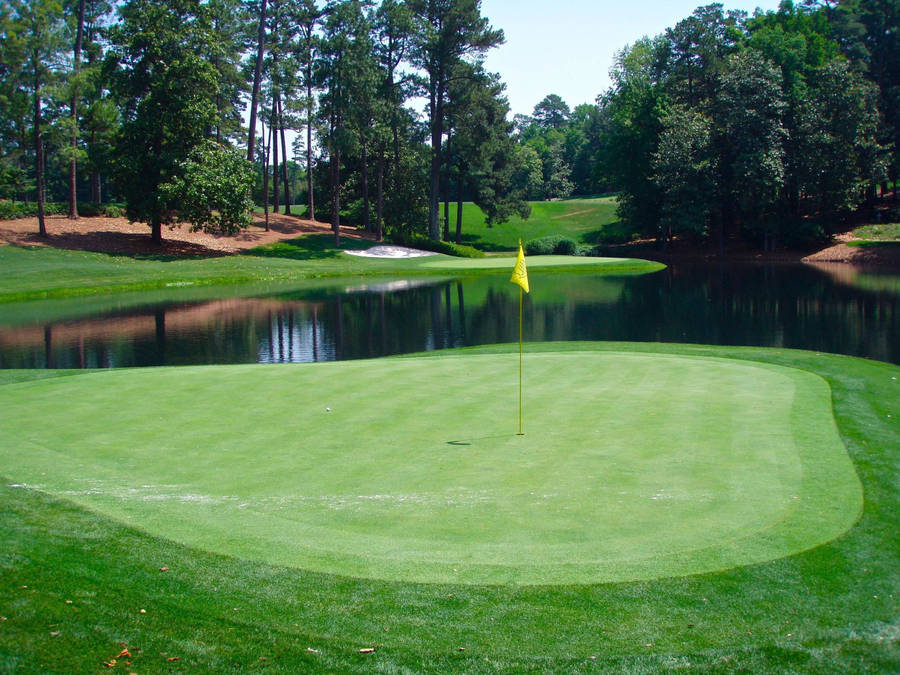 Green de golfe com bandeira amarela rodeado por água e árvores em um dia ensolarado. — Golfe Papéis de Parede