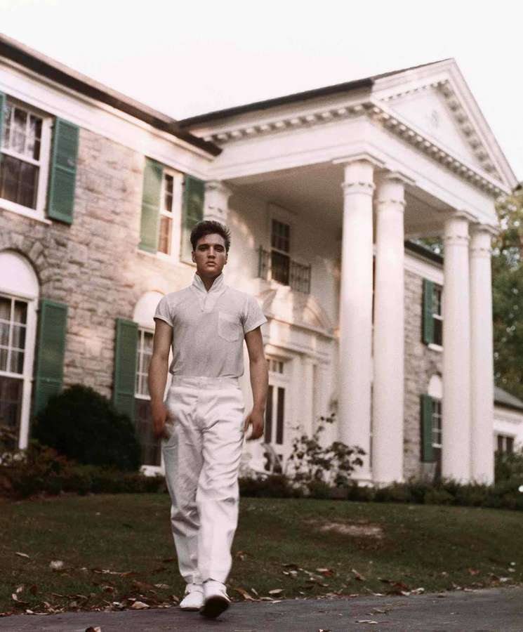 Young man in white outfit walking toward camera in front of large stone house with columns. — Graceland Wallpaper
