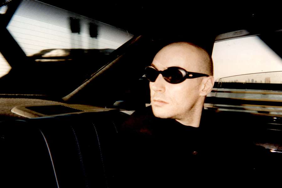 Bald man wearing sunglasses seated inside a vintage car with city view through window. — Grant Morrison Wallpaper