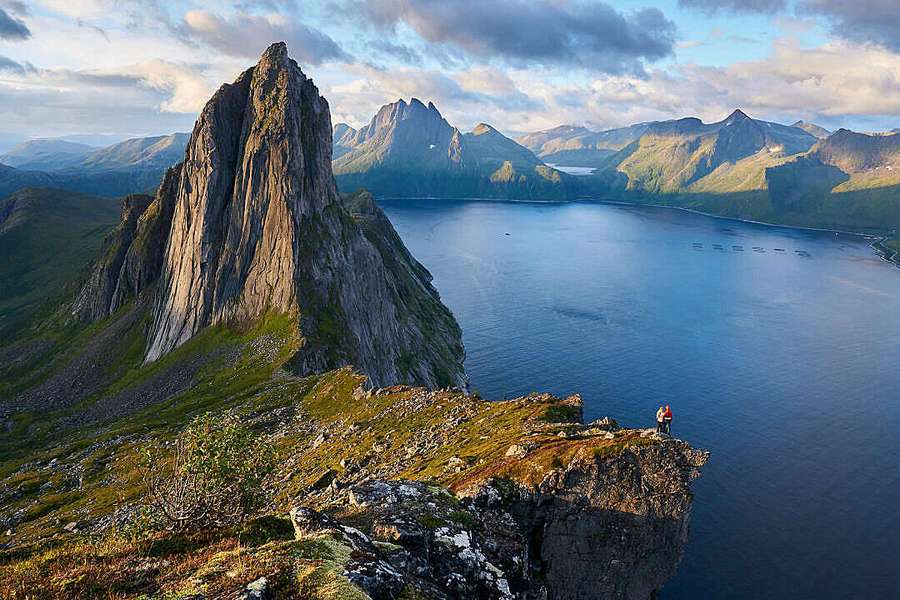 Eine Person steht auf einer Klippe mit Blick auf einen Fjord mit Bergen im Hintergrund. — HD Berg Wallpaper
