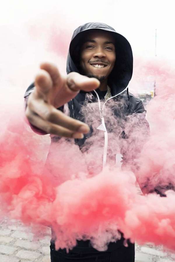 Homem de moletom sorrindo e alcançando através de fumaça rosa na calçada da cidade — Herbo iPhone Papéis de Parede