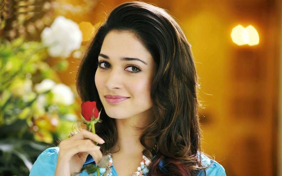 Woman holding red rose smiling in warm indoor setting with blurred flowers. — Heroine Background