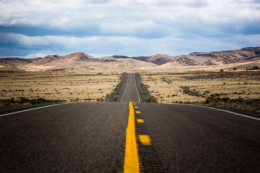 An empty road with yellow centerline leading through a dry landscape to distant hills. — Highway Picture