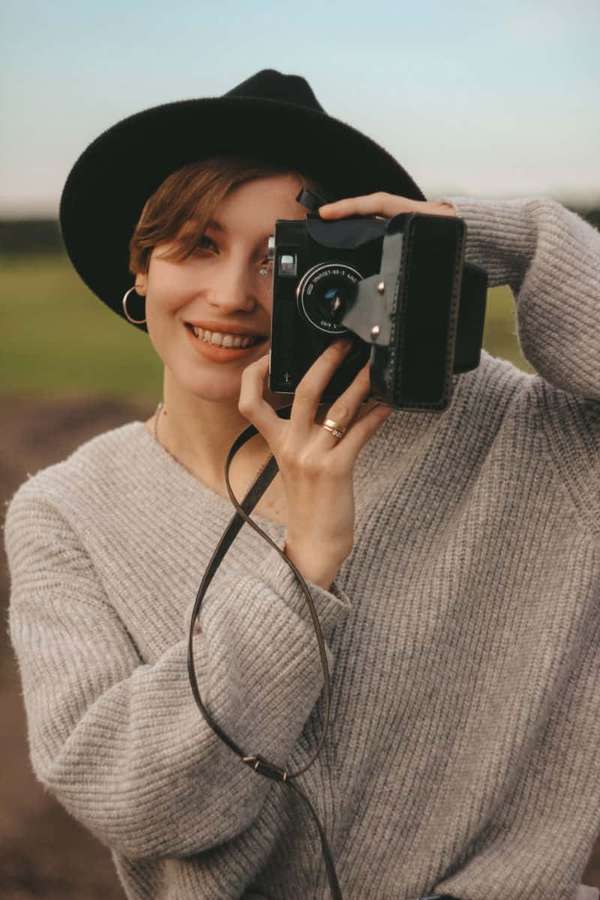 Woman in black hat and gray sweater holding vintage camera outdoors — Holding Camera Picture