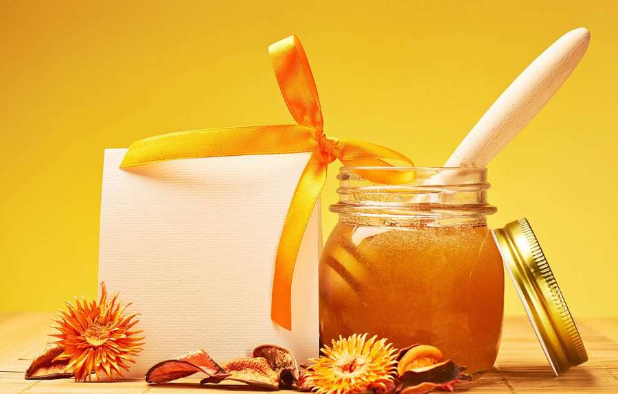 A jar of honey with a dipper next to a gift box and dried flowers on wood. — Honey Wallpaper