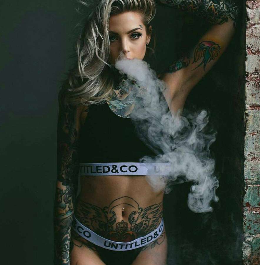 Tattooed woman exhaling smoke in branded underwear against a dark wall. — Hot Girls Wallpaper