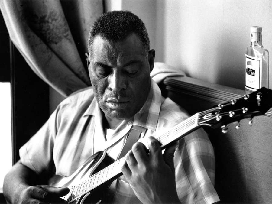 Man playing electric guitar in a dimly lit room with a bottle nearby. — Howlin Wolf Wallpaper