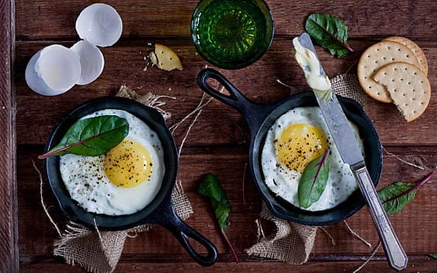 Dos huevos fritos con espinacas y pimienta en pequeñas sartenes sobre una mesa de madera con galletas y un vaso verde. — Huevos Fondos de pantalla