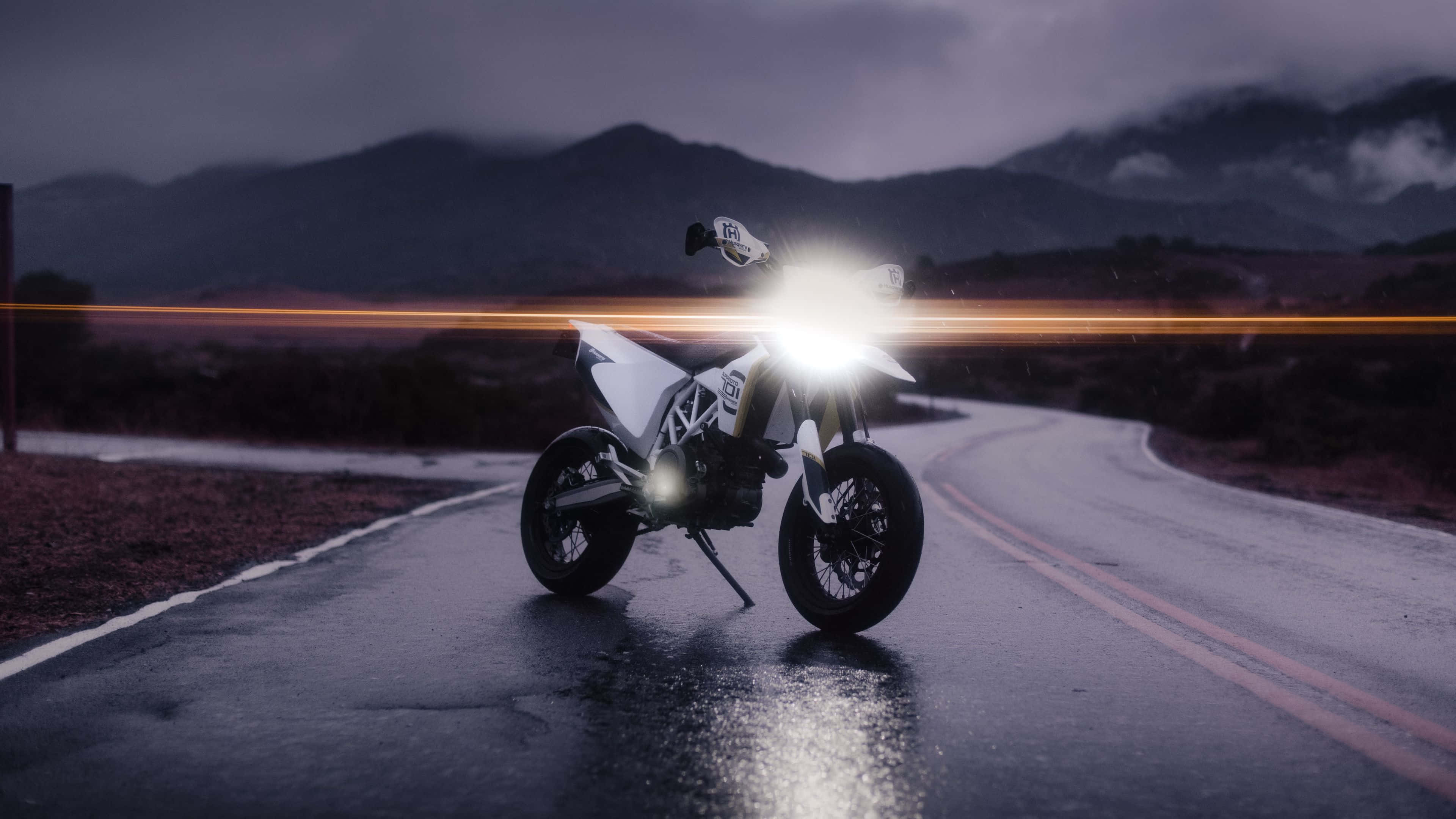 Hvid motorcykel på våd vej med skinnende forlygter under stormfuld himmel. — Husqvarna Wallpapers