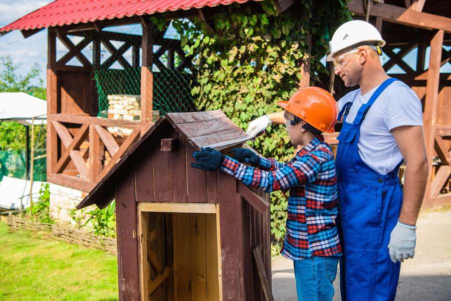 Padre e hijo usando cascos construyendo juntos una casa de madera para jugar al aire libre. — Imágenes de Casa de Perro Imágenes