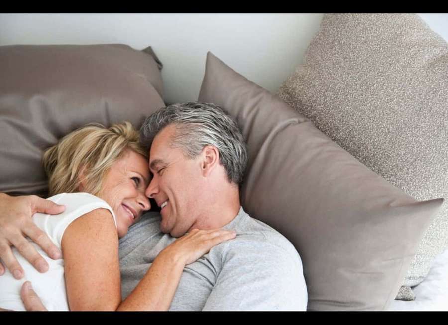 Um casal feliz se aconchegando em uma cama com travesseiros cinza. — Imagens de abraços Fotos