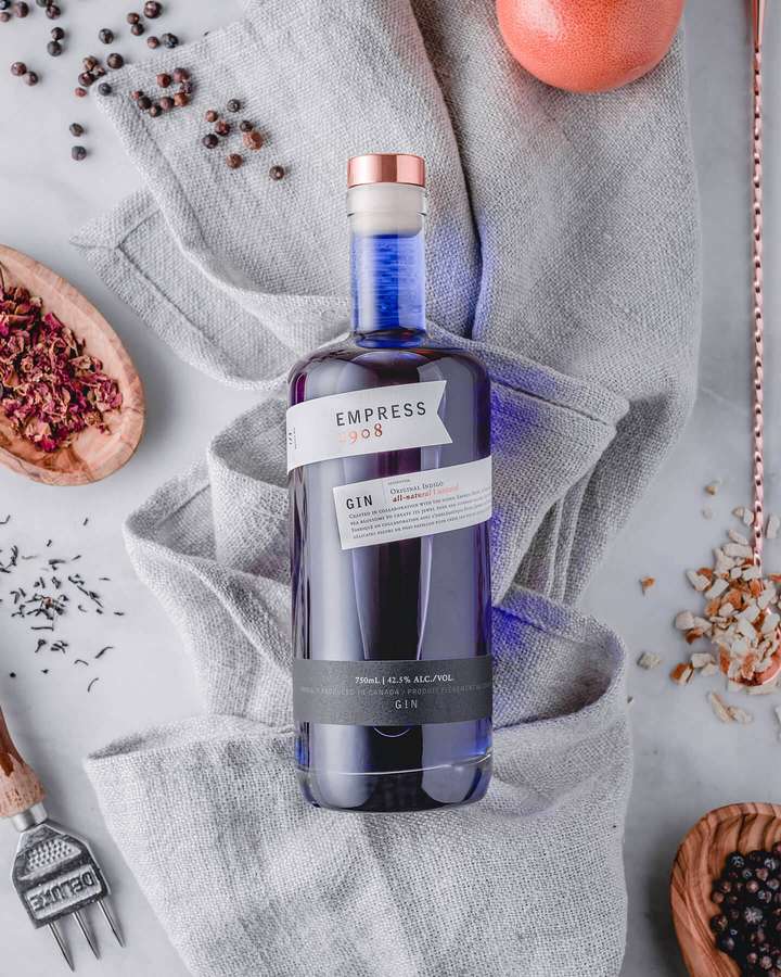 Uma garrafa de gin azul rodeada por ervas secas e frutas cítricas sobre um pano cinza. — Imperatriz Papéis de Parede