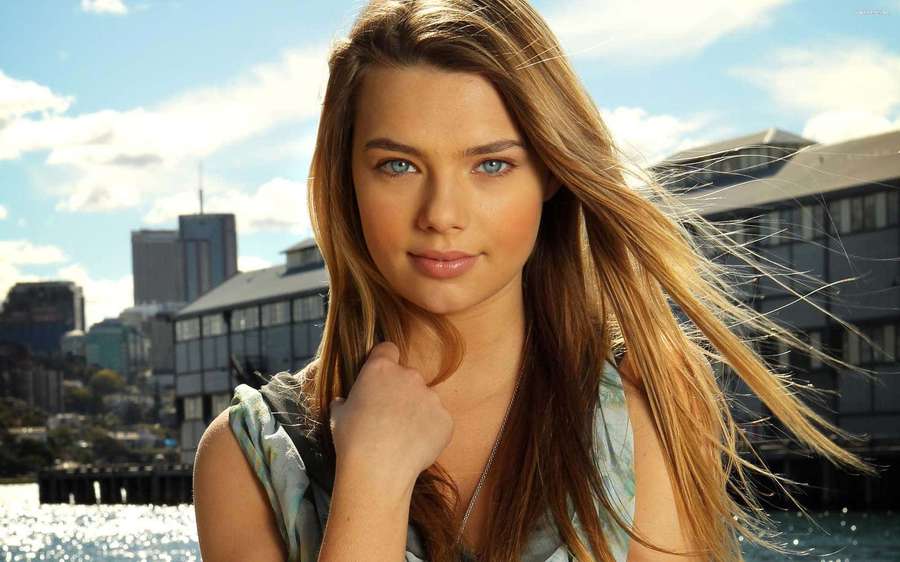 Young woman with blonde hair and blue eyes standing by water with city buildings behind her. — Indiana evans Wallpaper