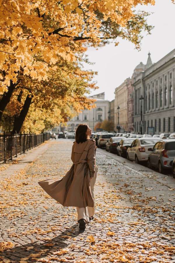 Woman in beige coat walking on leaf-covered cobblestone street in autumn city. — iPhone 11 Pro 4K Wallpaper