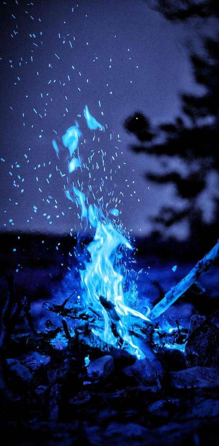 Blue campfire glowing at night with sparks flying and trees in silhouette. — iPhone Fire Picture