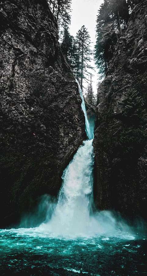 A turquoise waterfall plunges into a pool between dark forested cliffs. — iPhone Waterfall Wallpaper