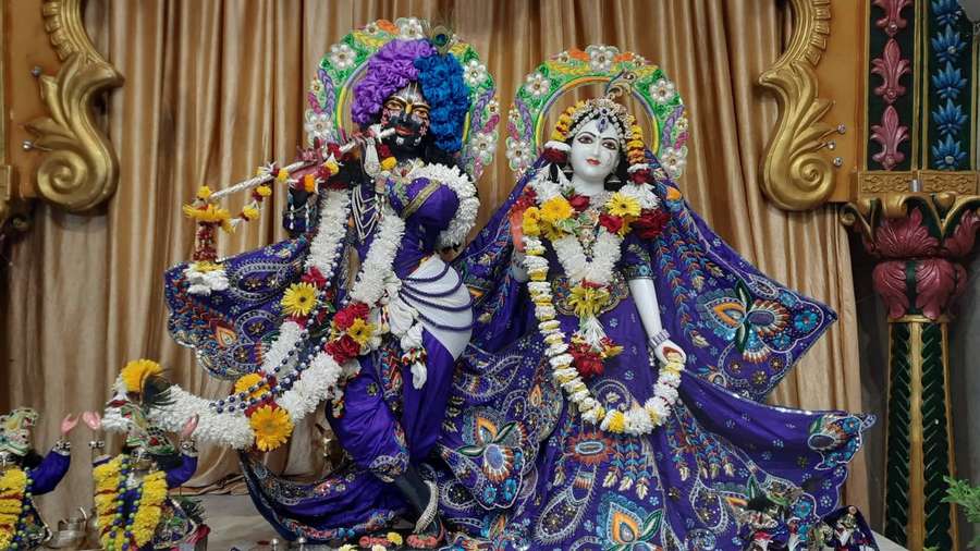 Zwei geschmückte hinduistische Gottheiten sitzen in einem verzierten Tempelumfeld mit Blumengirlanden. — Iskcon Wallpaper