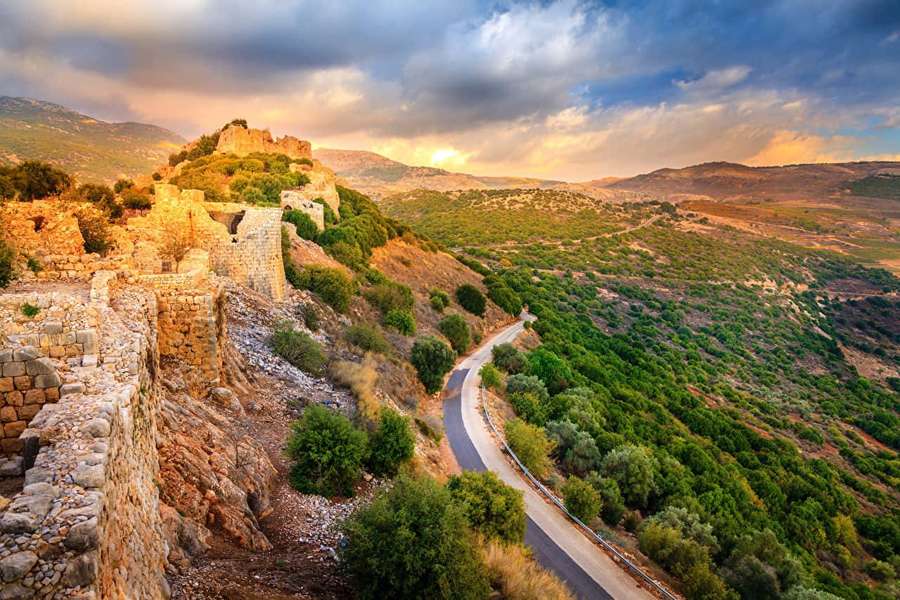 Ancient stone ruins overlook a winding road through green hills at sunset. — Israel Background