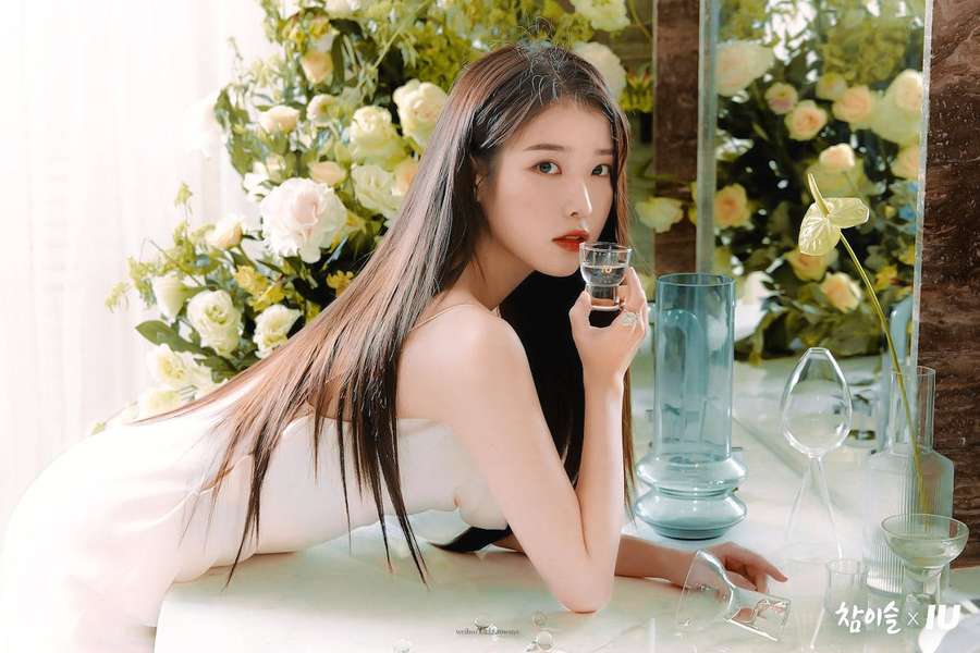 Woman in white dress holding glass surrounded by flowers and glassware. — Iu Background