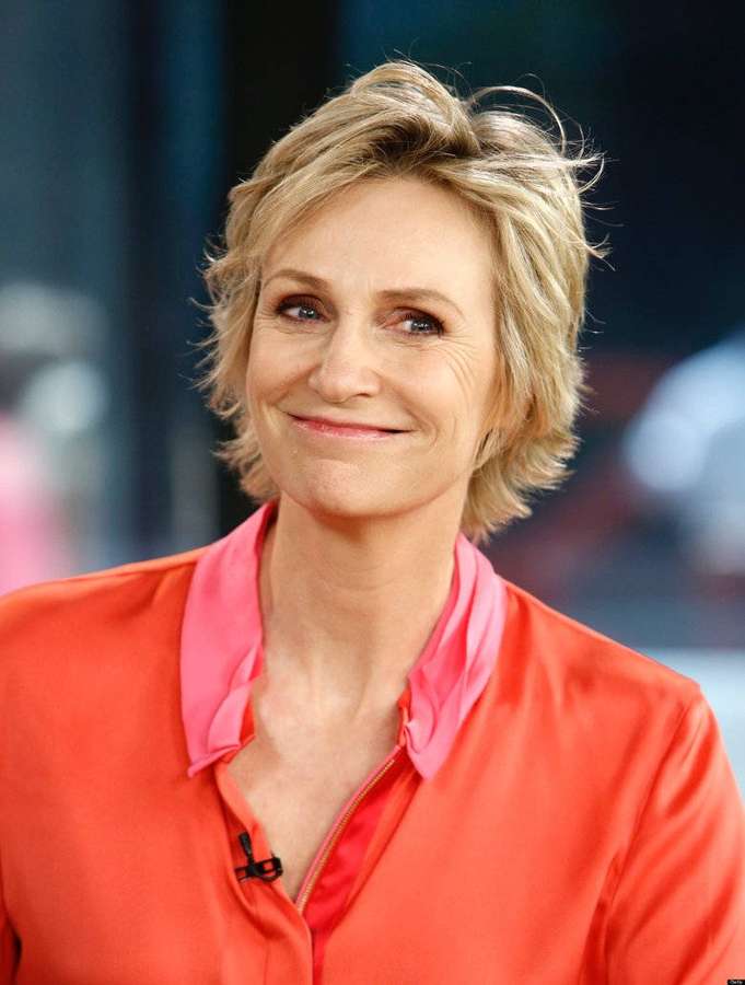 Woman with short blonde hair smiling in a coral blouse with pink collar. — Jane Lynch Wallpaper