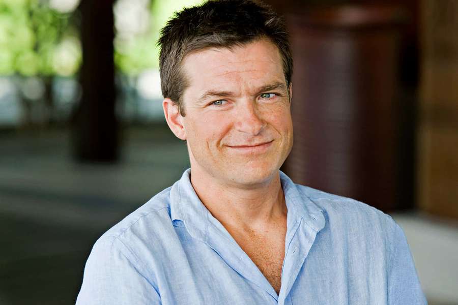 Un hombre con camisa azul claro sonríe al aire libre con árboles y columnas al fondo. — Jason Bateman Fondos de pantalla