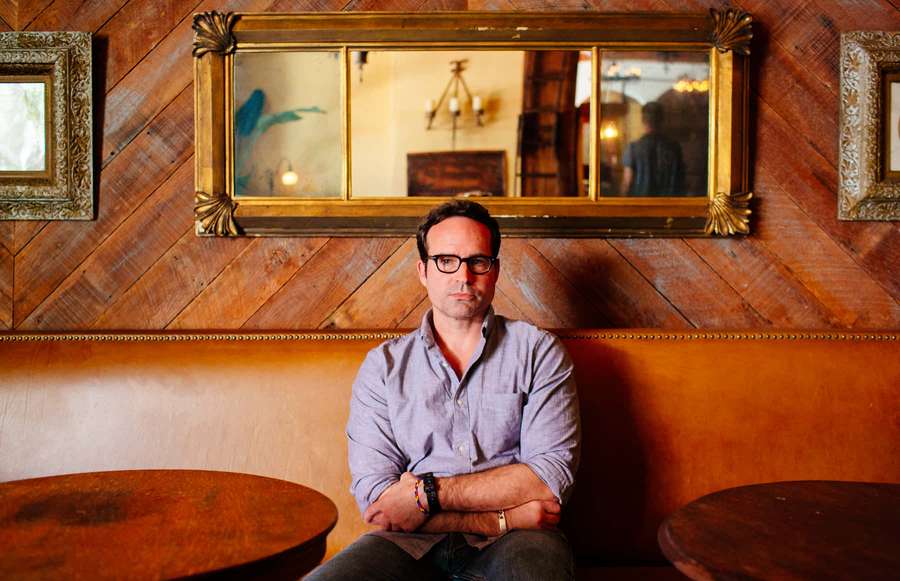 Man with glasses sitting on a leather booth in a wooden room with mirrors. — Jason Patric Wallpaper