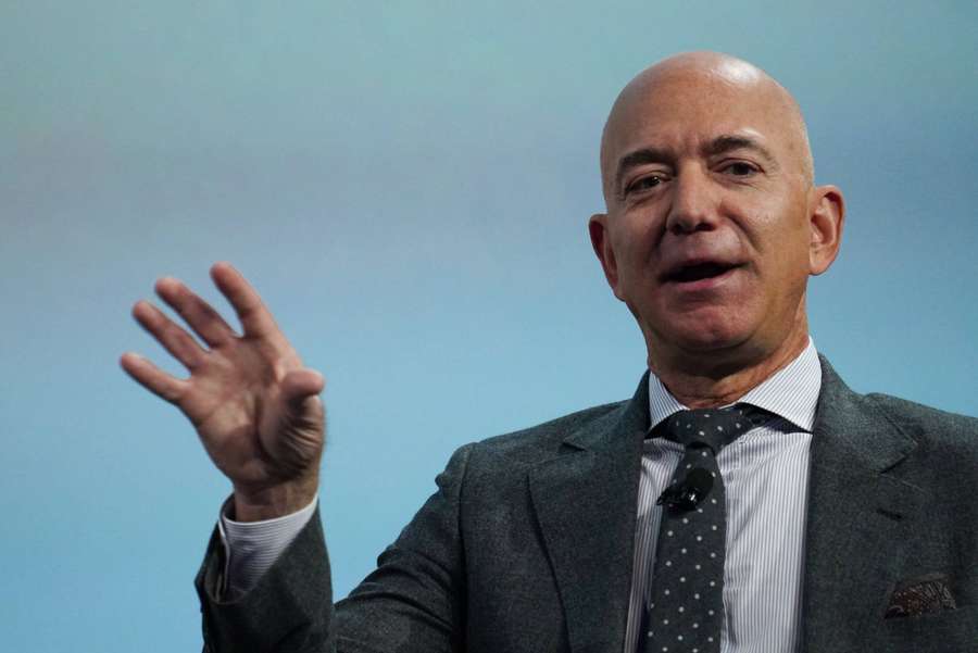 Jeff Bezos speaking in a suit with raised hand against blue background. — Jeff Background