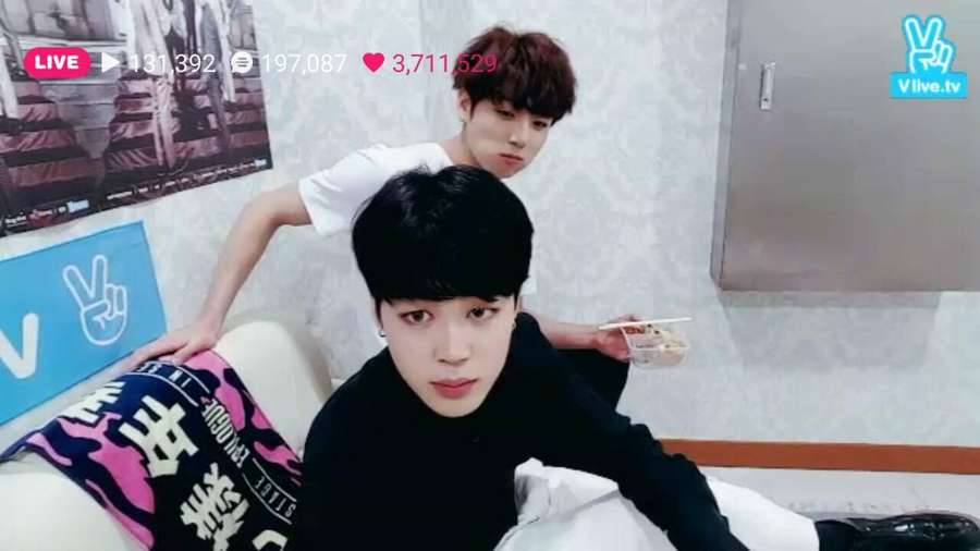 Zwei junge Männer in einem Raum, einer sitzt auf einem Sofa und hält Essstäbchen, der andere lehnt sich hinter ihn, beide schauen in die Kamera mit Live-Stream-Overlays. — Jikook Bilder
