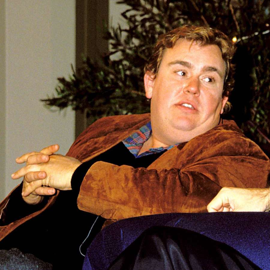 Man in brown velvet robe sitting indoors with clasped hands — John Candy Wallpaper