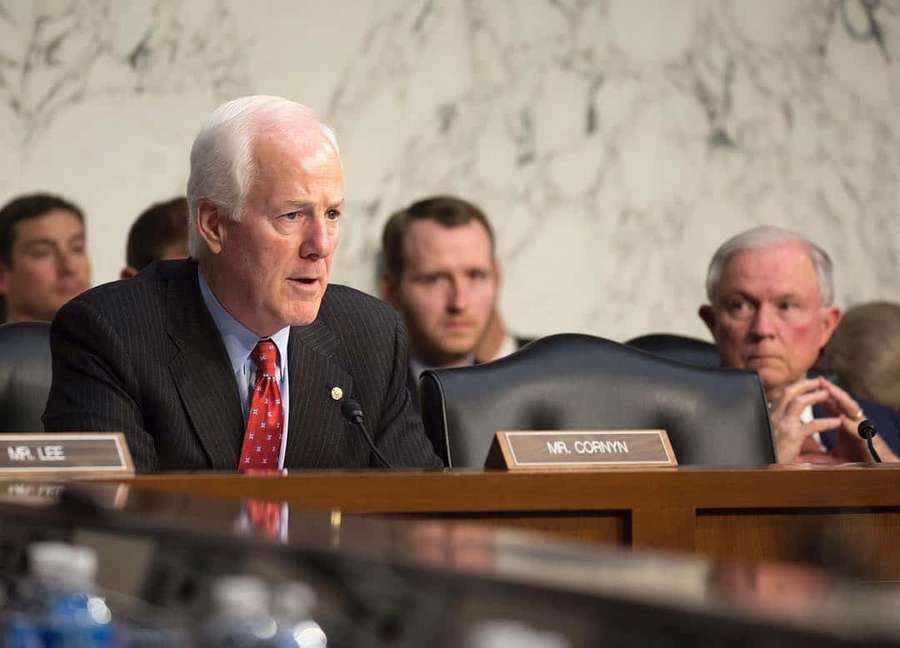 Pria bersetelan berbicara di sidang pemerintah dengan papan nama dan mikrofon. — John Cornyn Wallpaper