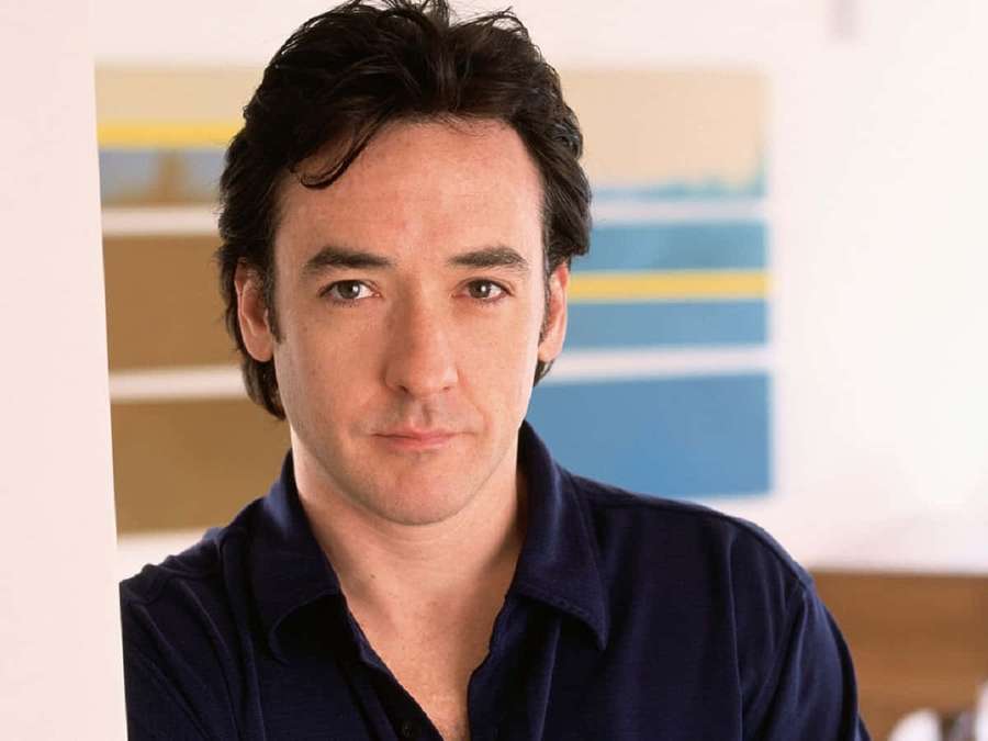 A man in a navy blue shirt looks calmly at the camera indoors. — John Cusack Wallpaper