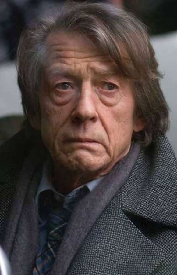 Ein älterer Mann mit grauen Haaren, der einen Mantel und Schal trägt, blickt ernst nach vorne. — John Hurt Bilder