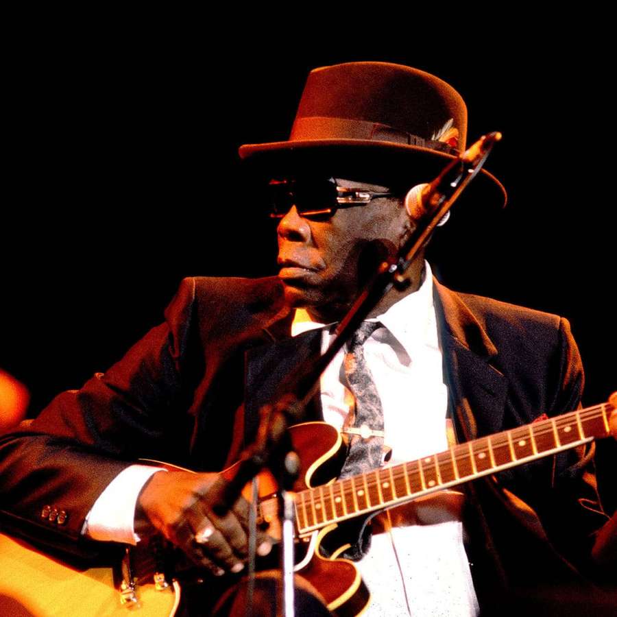 A man in a hat and sunglasses plays an electric guitar on stage. — John Lee Hooker Wallpaper