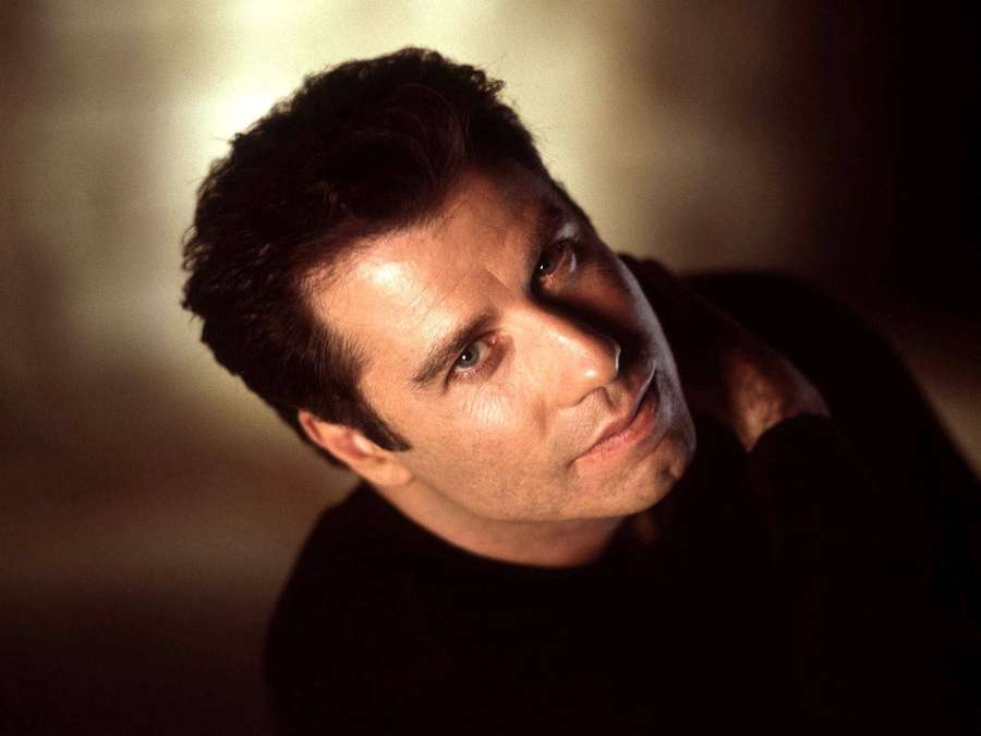 A man in a dark shirt looks upward with a serious expression in warm studio lighting. — John Travolta Wallpaper