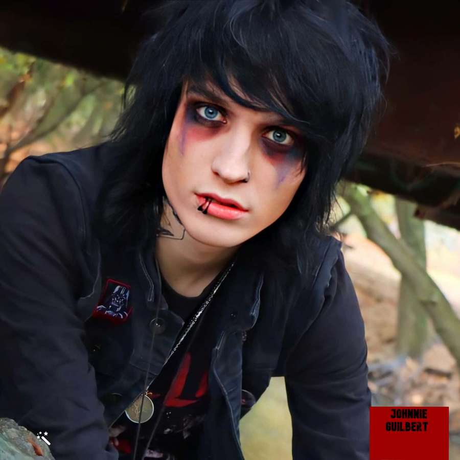Henkilö, jolla on goottimeikki ja mustat hiukset, poseeraa ulkona puurakenteen alla. — Johnnie Guilbert Taustakuvat
