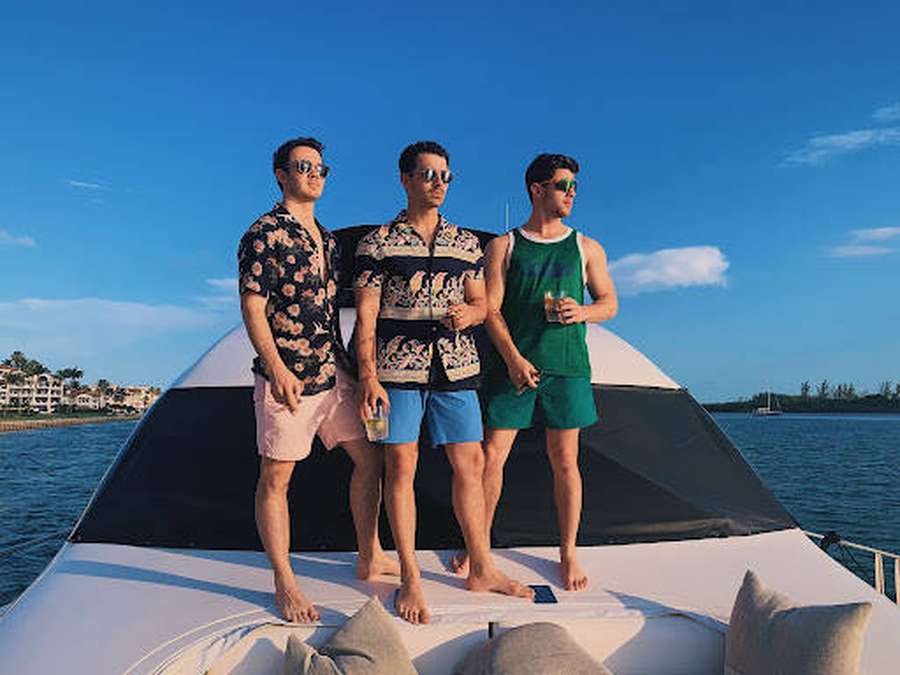 Three men in aloha shirts on a yacht enjoying drinks at sunset. — Jonas Brothers Wallpaper