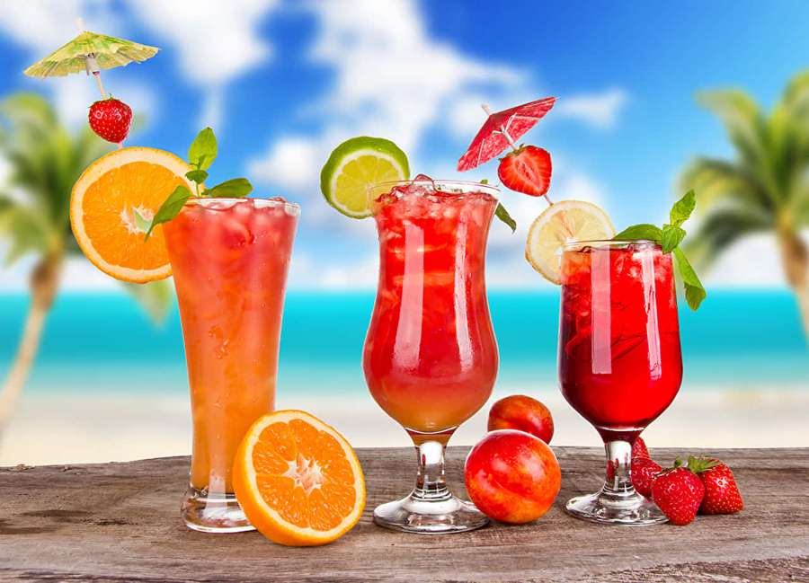 Three colorful tropical drinks with fruit garnishes on a beach table. — Juice Wallpaper